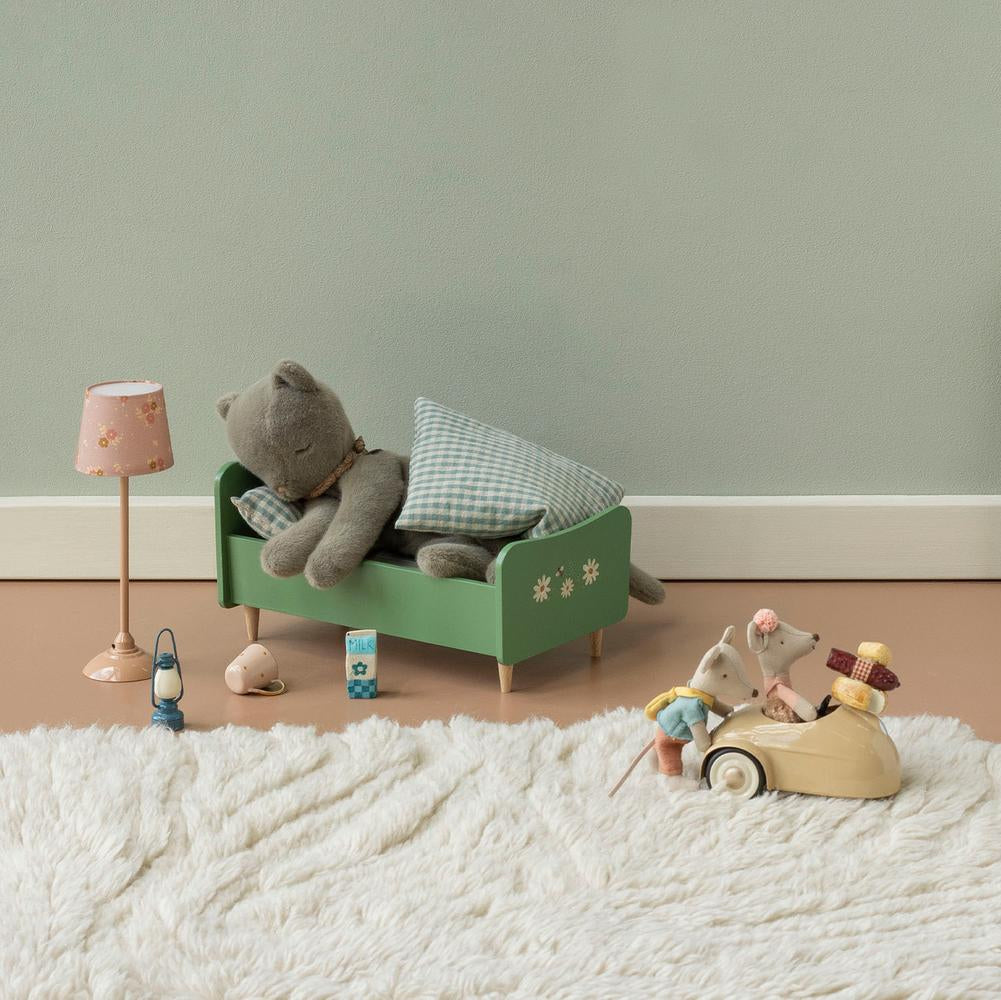 A styled children's bedroom scene with a green wooden bed frame holding a gray stuffed bear and pillows, a pink lamp, a small green cabinet, and a wooden mouse toy driving a car on a white shag rug against a pale green wall.