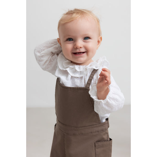 Child wearing a white ruffled shirt and brown overalls on a light background