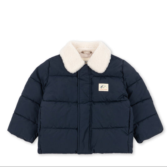 A navy blue puffer jacket with a cream-colored faux fur collar, featuring horizontal quilting and a small rectangular brand patch on the left chest, displayed against a solid white background.