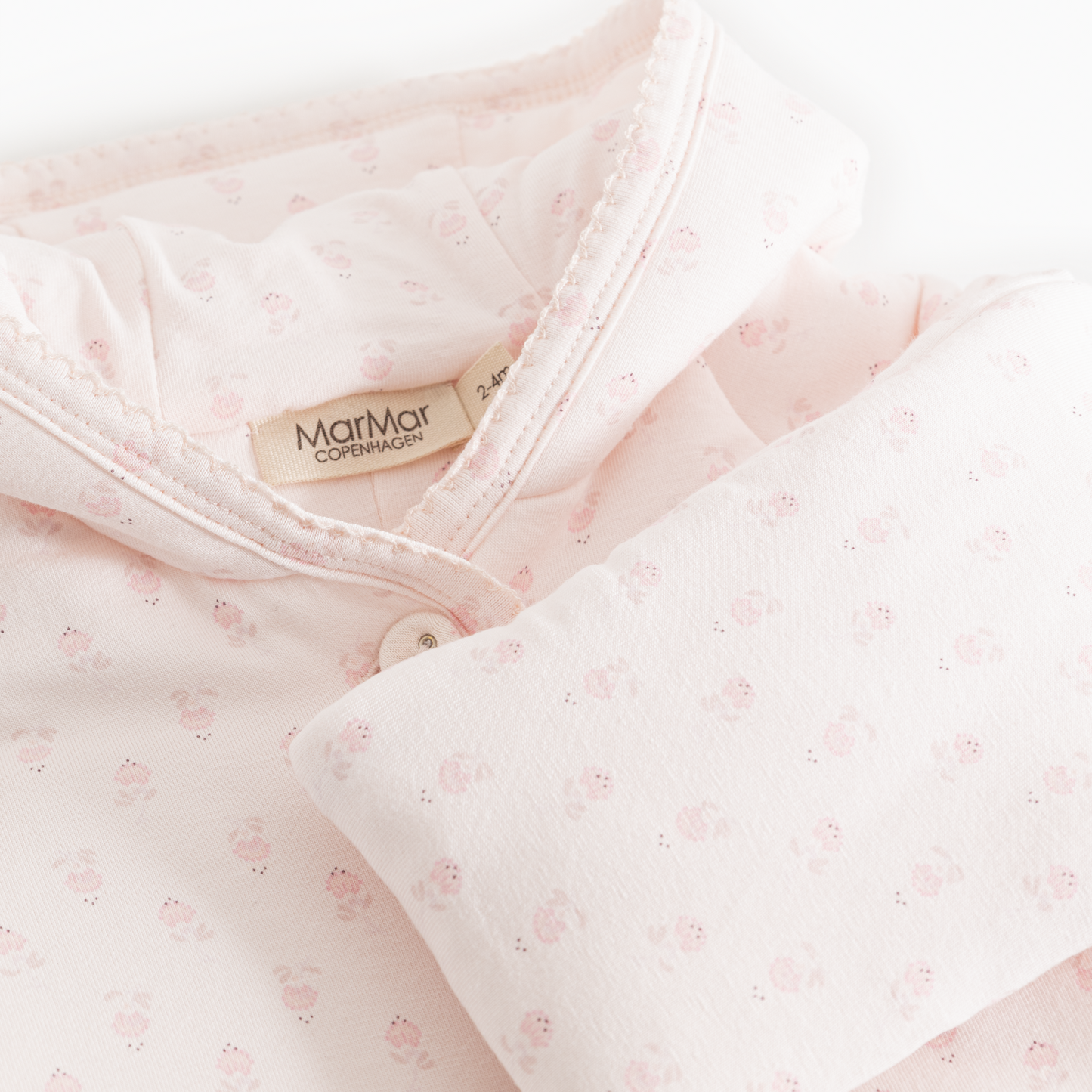 Close-up of a light pink baby garment with a small allover floral print, featuring a soft collar, button closure, and a visible MarMar Comfortcare label.