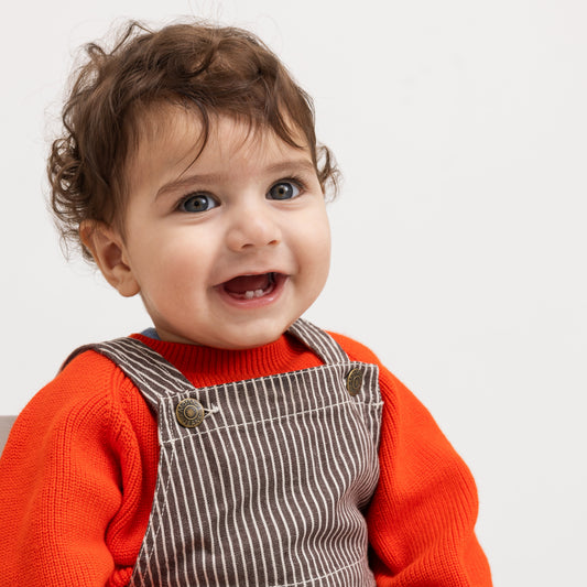 A close-up of a smiling toddler with curly brown hair, wearing a bright orange knit sweater and brown corduroy-style overalls with thin white stripes, photographed against a plain light background.