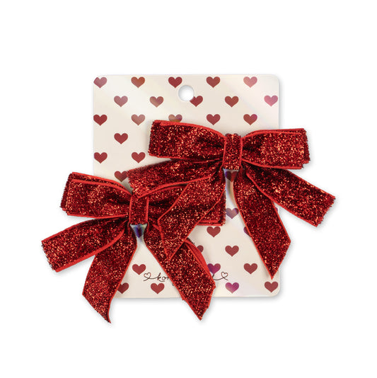 A set of two red glitter hair bows attached to a white display card decorated with small red hearts, photographed on a plain white background.