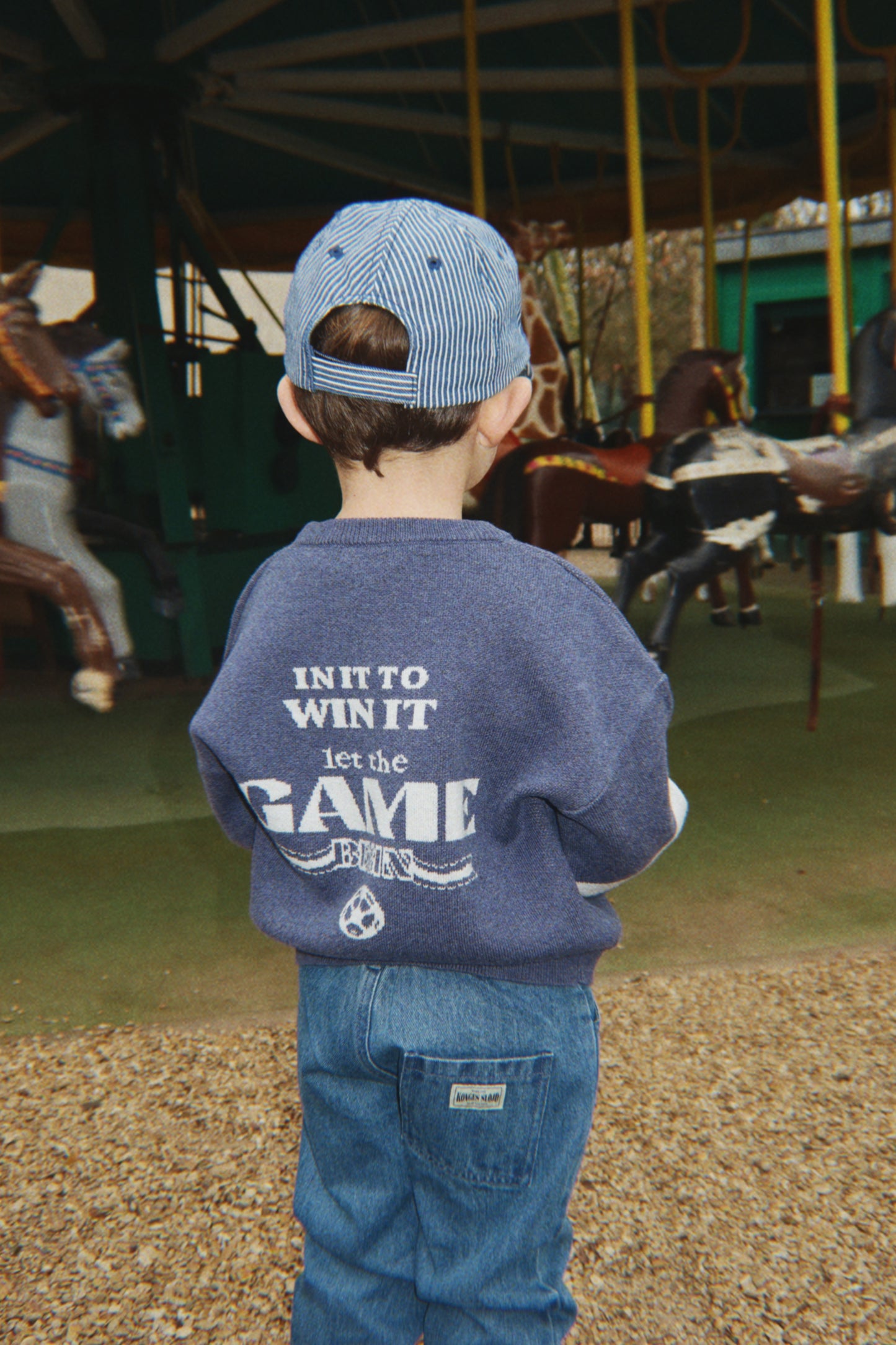 Child wearing a blue sweatshirt with text and cap, standing in an indoor equestrian arena.