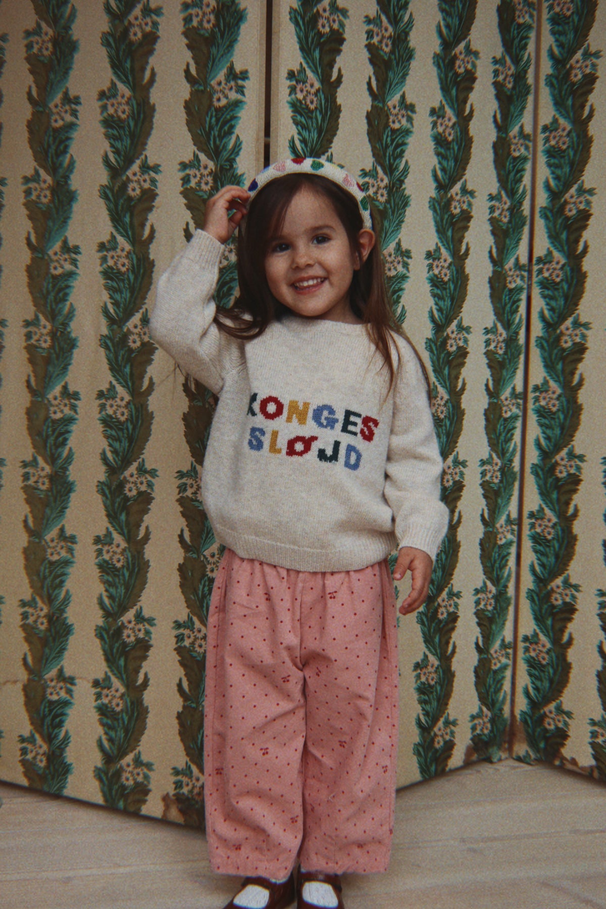 A young girl smiling and posing in front of a vintage patterned wallpaper background. She is wearing a cream-colored knit sweater with colorful embroidered text reading 'KONGES SLOJOD', pink patterned trousers, and a matching pink and white polka dot headband.