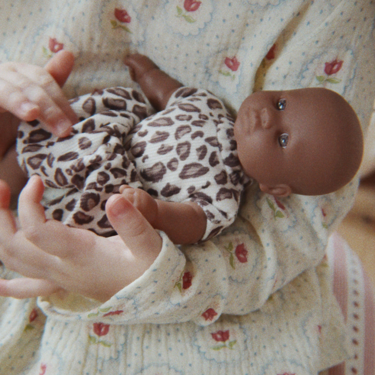 A close-up lifestyle shot of a person holding a baby doll. The person is wearing a cream-colored long-sleeve knit top with a red and green floral pattern, and the doll is dressed in a brown and white leopard print outfit.