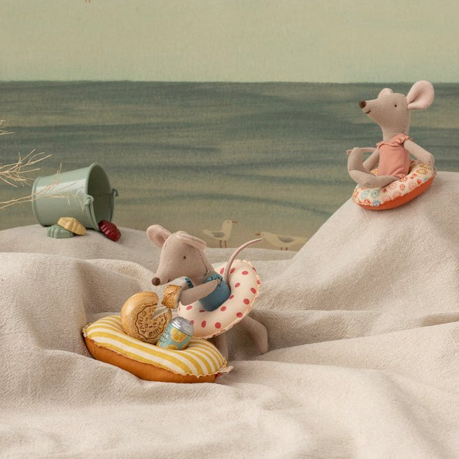 Two toy mice with beach accessories on a sandy surface with a scenic background.