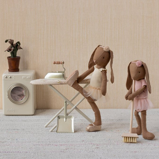 Two toy rabbit figures in a domestic setting with a washing machine, ironing board, and broom.