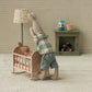 Stuffed rabbit in plaid shirt and shorts standing next to a baby crib in a room with a lamp and fireplace.