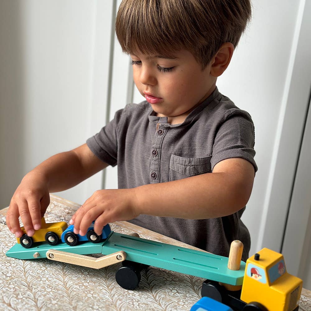 A young boy with brown hair wearing a gray short-sleeve henley shirt is playing with a wooden toy truck and trailer set on a patterned tablecloth.