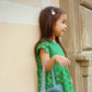 Young girl in a green dress with cherry design leaning against a stone wall.