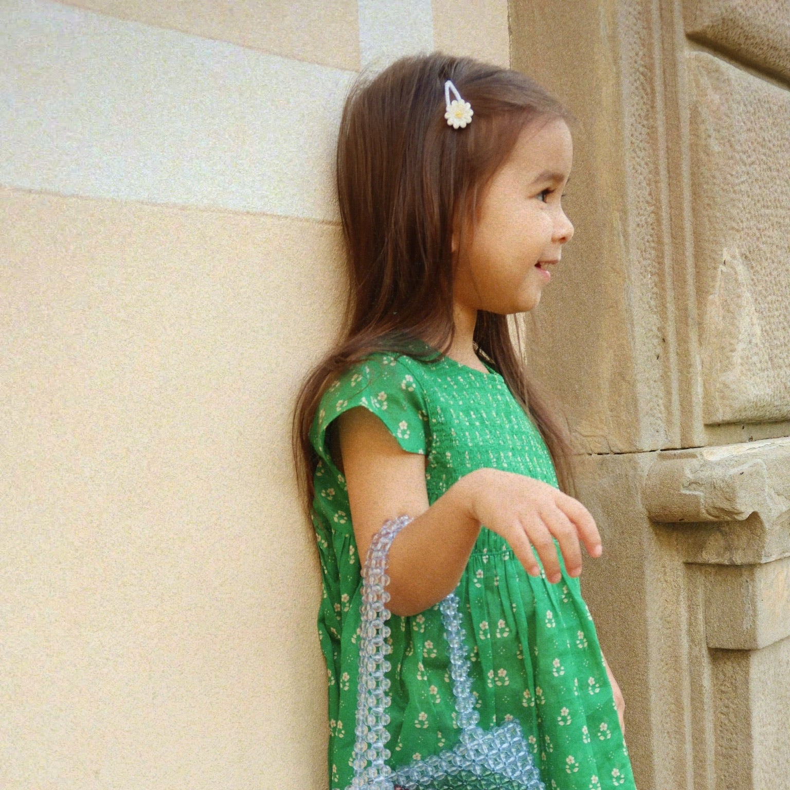 Young girl in a green dress with cherry design leaning against a stone wall.