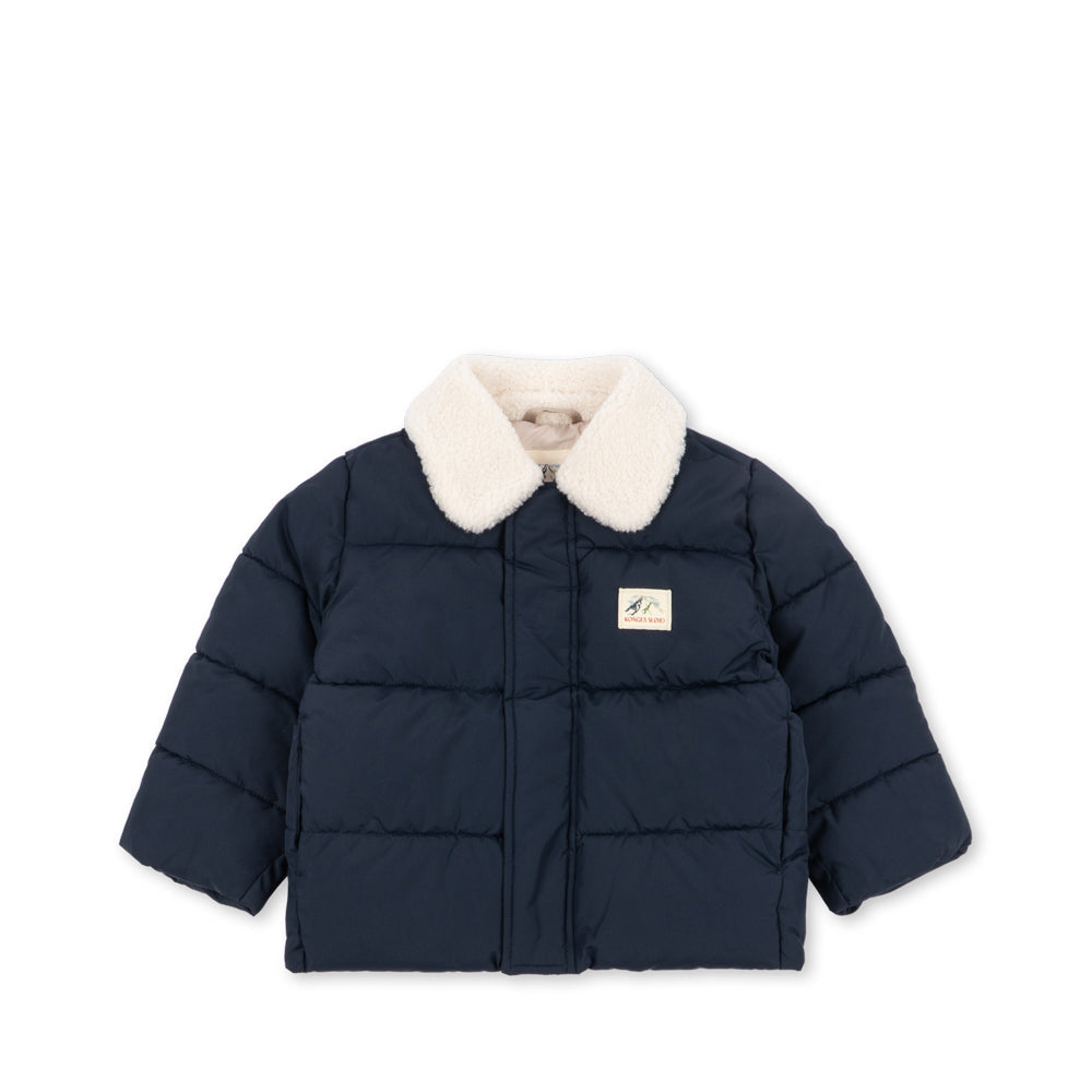 A navy blue puffer jacket with a cream-colored faux fur collar, featuring horizontal quilting and a small rectangular brand patch on the left chest, displayed against a solid white background.