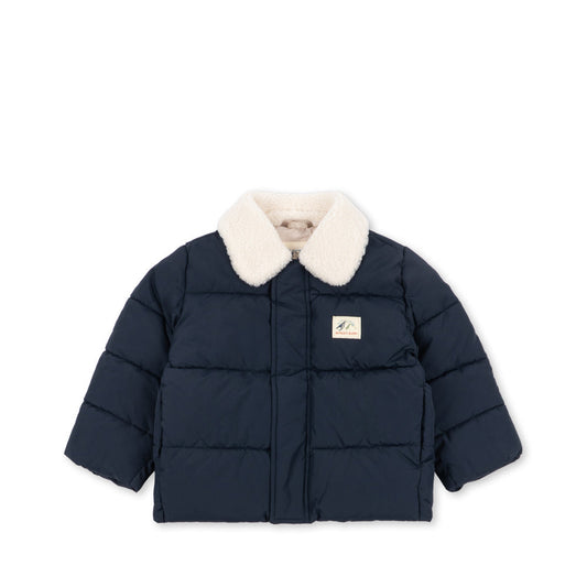 A navy blue puffer jacket with a cream-colored faux fur collar, featuring horizontal quilting and a small rectangular brand patch on the left chest, displayed against a solid white background.