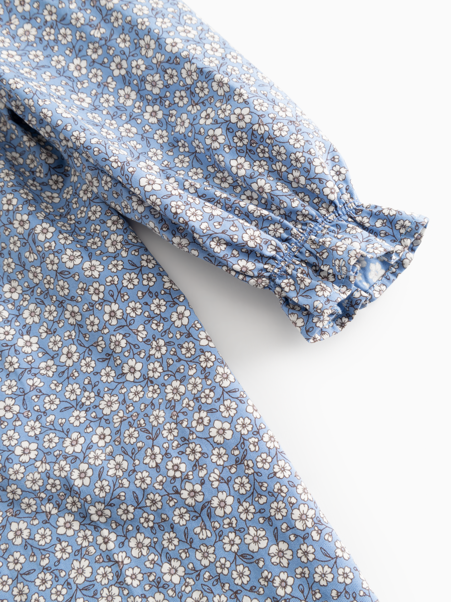 Close-up of a blue fabric garment with a small white and beige floral print and a gathered elastic cuff on the sleeve, shown against a light gray background.