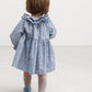 A toddler viewed from behind wearing a blue and white floral print dress with a ruffled collar, light pink knit socks, and blue shoes, standing against a plain white studio background.
