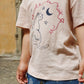 Girl wearing a beige t-shirt with a cat graphic and text, standing outdoors.