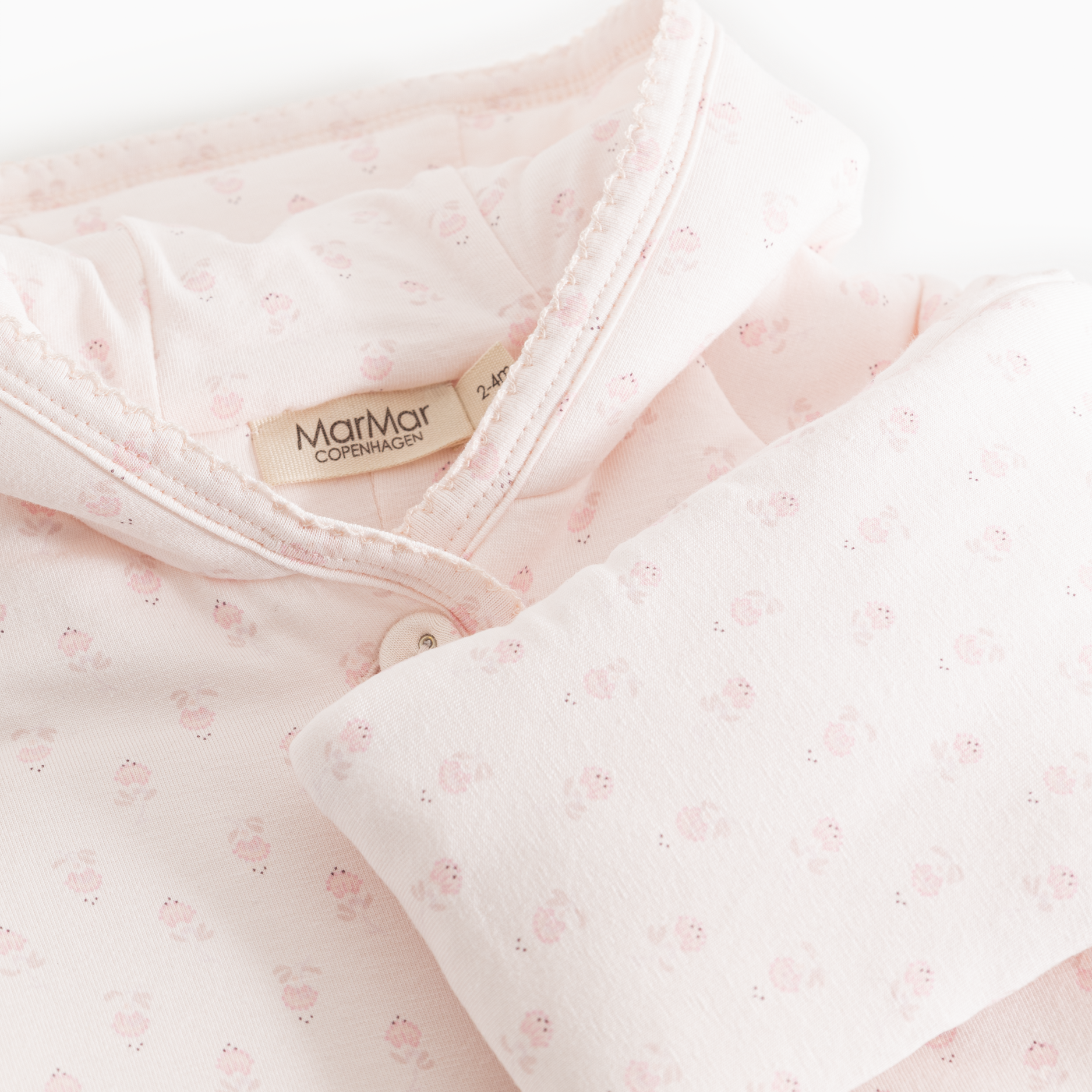 Close-up of a light pink baby garment with a small allover floral print, featuring a soft collar, button closure, and a visible MarMar Comfortcare label.