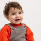 A close-up of a smiling toddler with curly brown hair, wearing a bright orange knit sweater and brown corduroy-style overalls with thin white stripes, photographed against a plain light background.