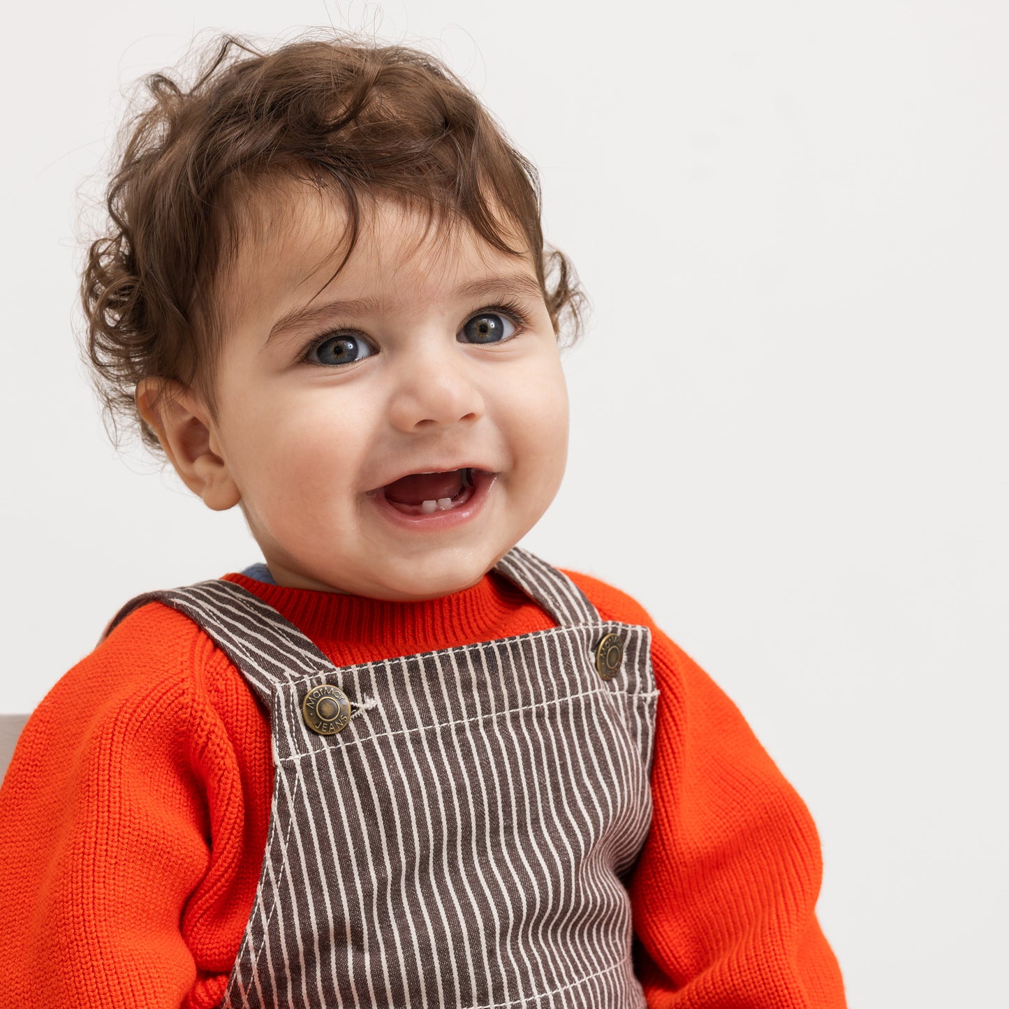 A close-up of a smiling toddler with curly brown hair, wearing a bright orange knit sweater and brown corduroy-style overalls with thin white stripes, photographed against a plain light background.