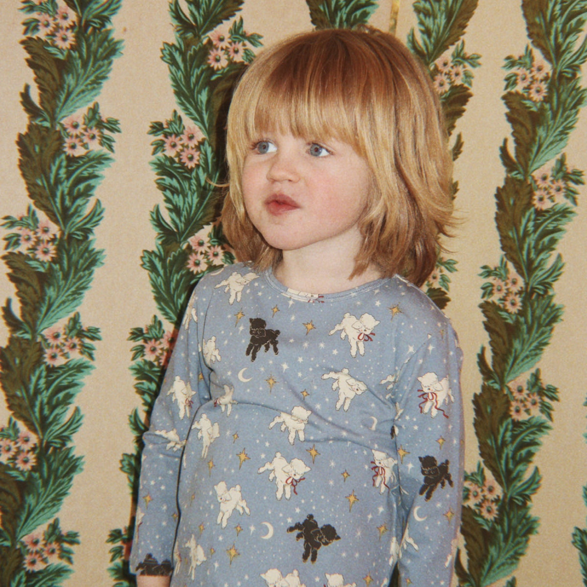 A young child with blonde hair wearing a light blue long-sleeve pajama set with a white and black unicorn pattern, standing in front of a vintage floral wallpaper background.