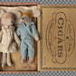 Two teddy bears, a girl in a dress and a boy in overalls, inside an open cigar box with vintage design.