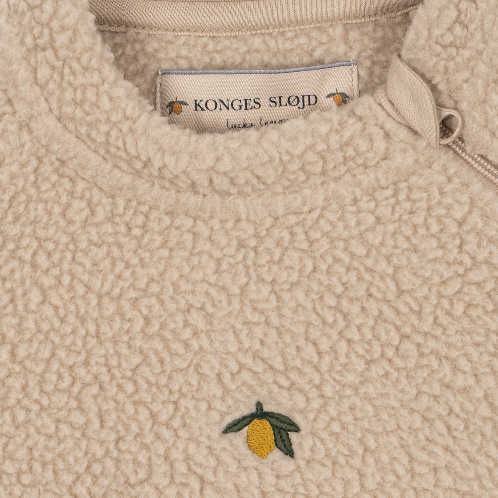 Close-up of a beige sherpa fleece zip-up sweater with a small embroidered yellow lemon and green leaf on the chest. The brand label 'KONGES SLOJ' is visible inside the collar.