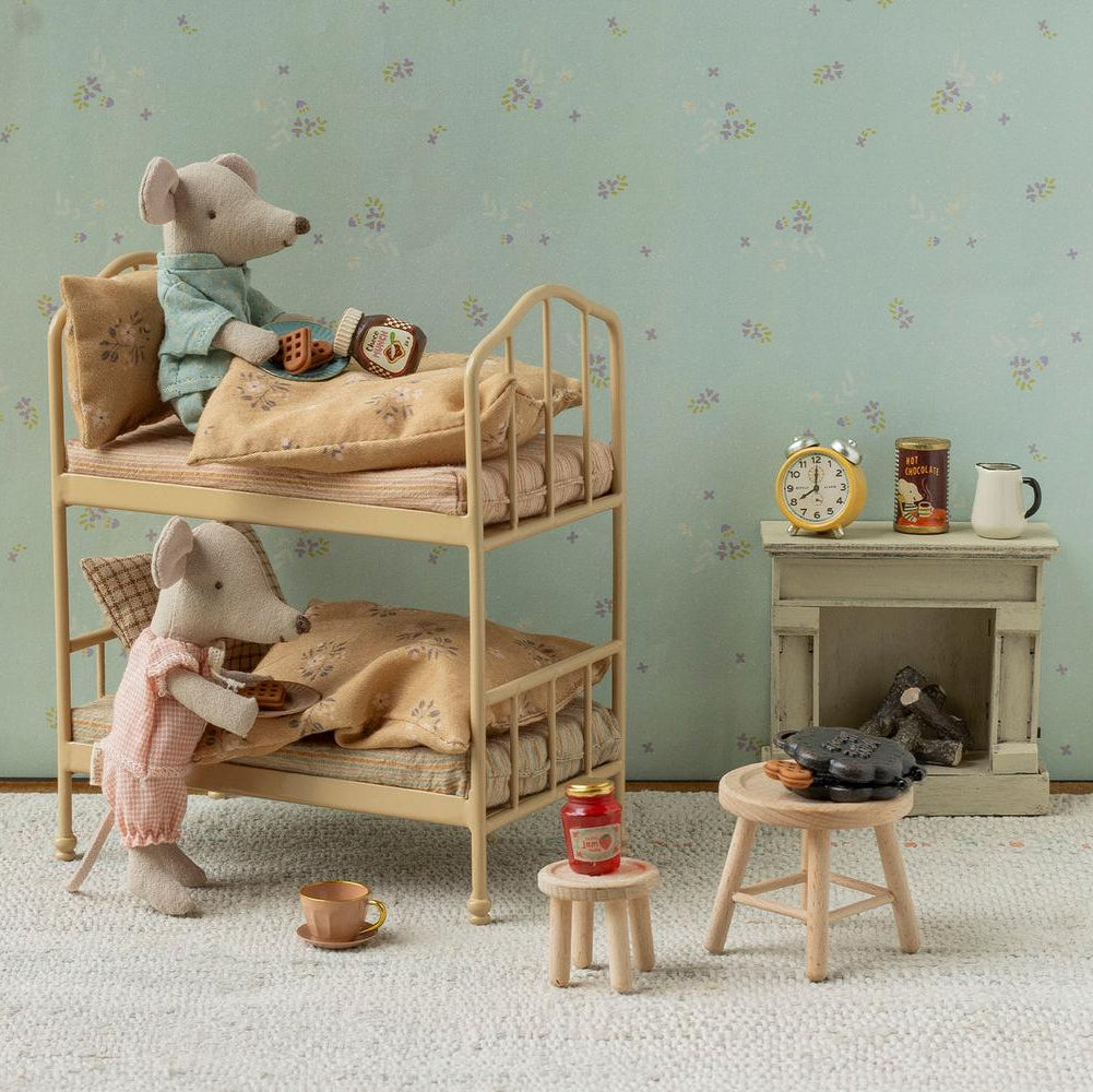 A miniature dollhouse-style room scene with two plush mouse figures arranged on a wooden bunk bed, alongside small furniture pieces, a wallpapered wall, and a fireplace display.