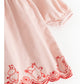 A close-up of a light pink fabric garment, likely a dress or nightgown, featuring red embroidered cartoon dog characters and a scalloped red border along the hem.