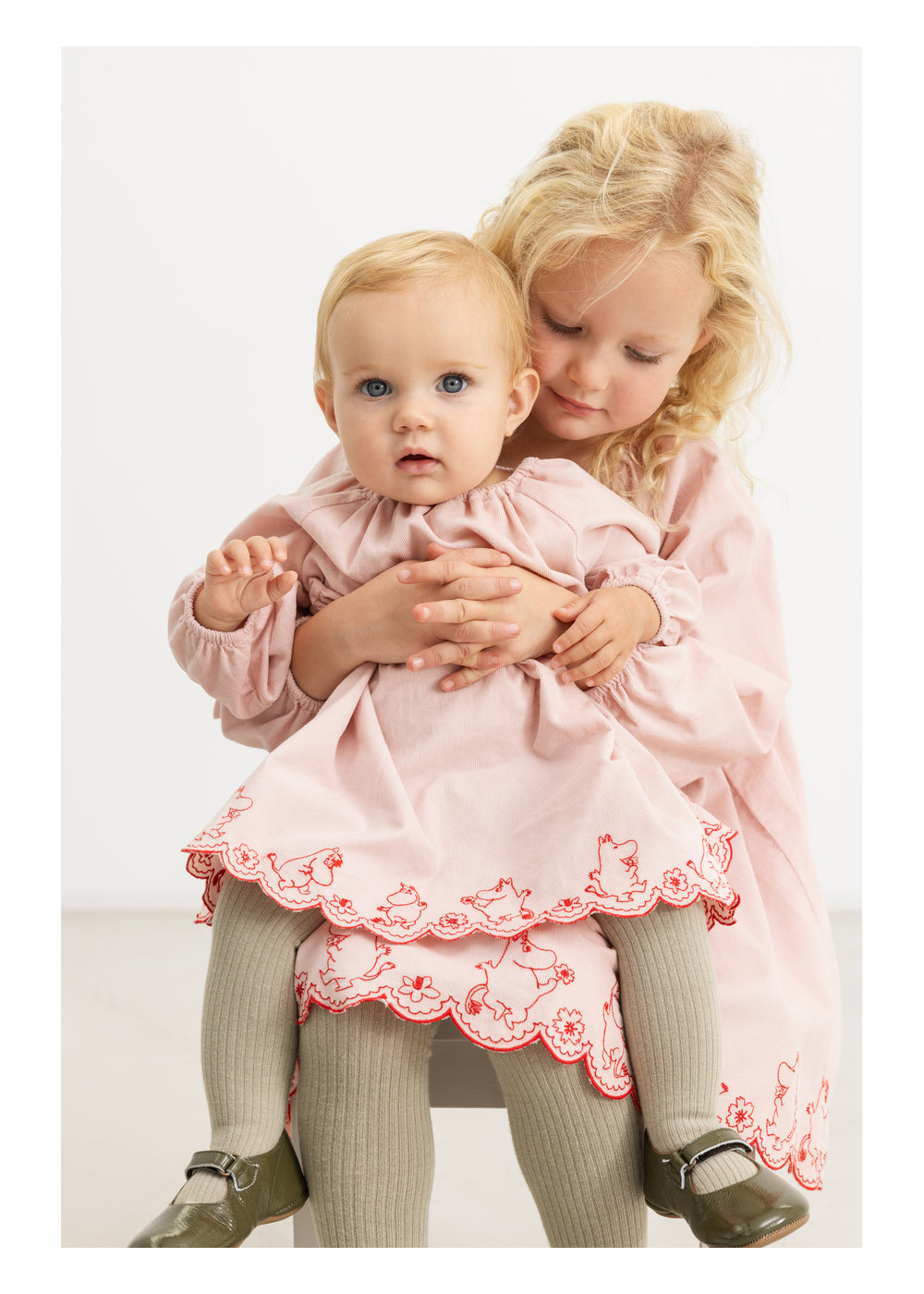 Two young girls in matching light pink dresses with red embroidered floral borders and gray tights, posed against a clean white studio background.