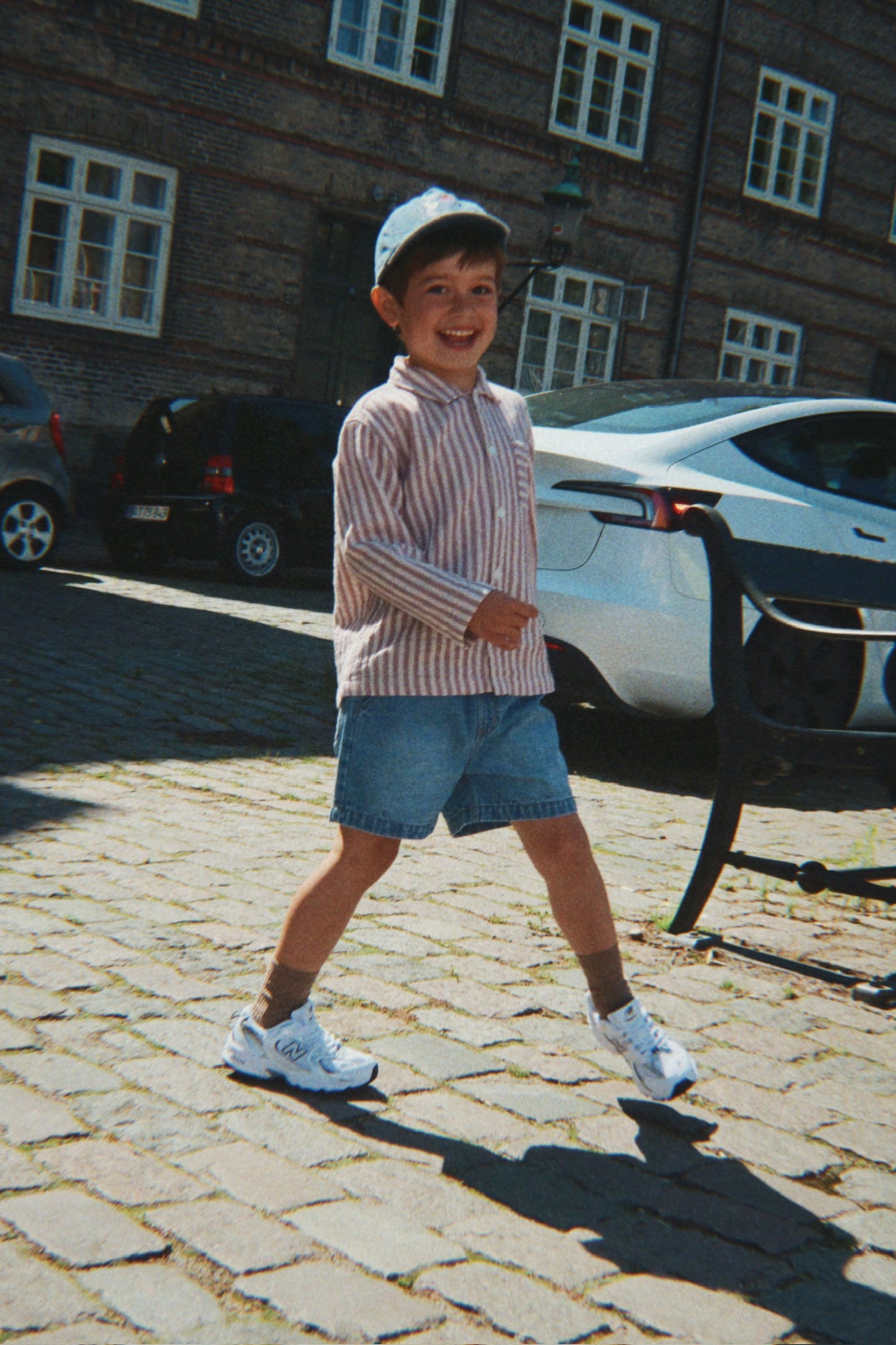 Child standing on a cobblestone street with a building and cars in the background