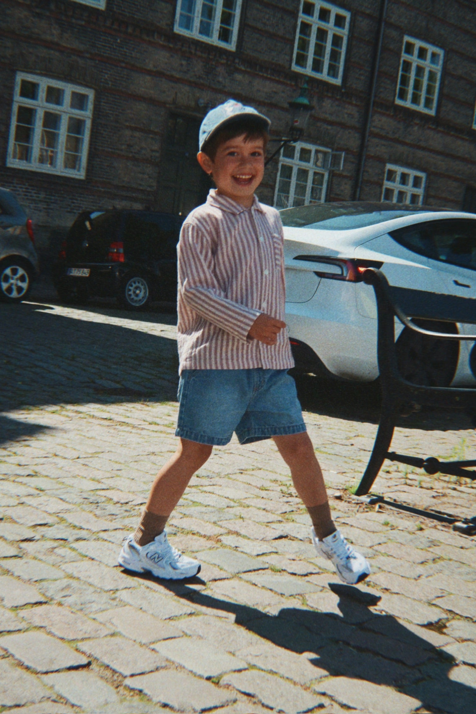 Child standing on a cobblestone street with a building and cars in the background
