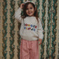 A young girl smiling and posing in front of a vintage patterned wallpaper background. She is wearing a cream-colored knit sweater with colorful embroidered text reading 'KONGES SLOJOD', pink patterned trousers, and a matching pink and white polka dot headband.