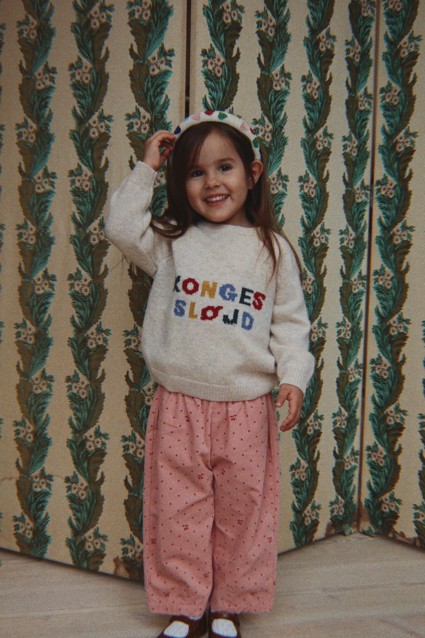 A young girl smiling and posing in front of a vintage patterned wallpaper background. She is wearing a cream-colored knit sweater with colorful embroidered text reading 'KONGES SLOJOD', pink patterned trousers, and a matching pink and white polka dot headband.