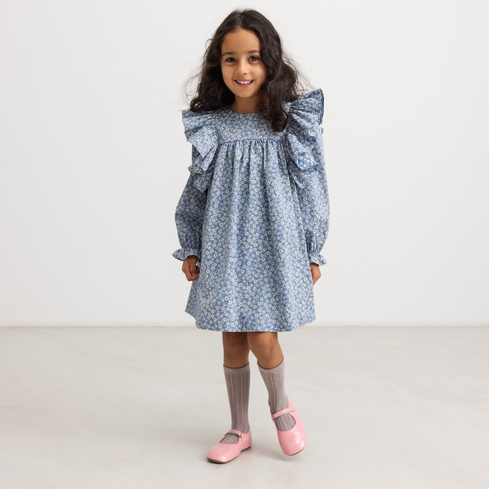 A young girl with long dark hair stands in a studio wearing a light blue and white floral print dress with dramatic ruffled shoulders, gray socks, and pink ballet flats.