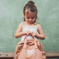 Young girl holding a brown backpack against a green wall.