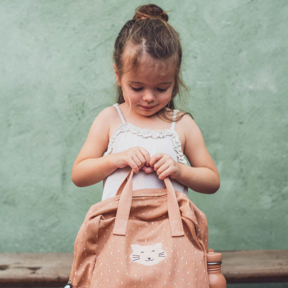Young girl holding a brown backpack against a green wall.