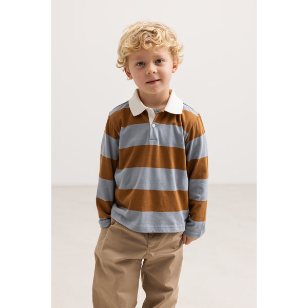 A young boy with curly blonde hair wearing a long-sleeve rugby shirt with brown and light gray horizontal stripes, paired with khaki pants, standing against a plain off-white studio background.