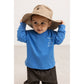A toddler wearing a bright blue long-sleeve shirt with a small white graphic on the chest, dark brown pants, and a beige wide-brim sun hat, posing against a light off-white background.
