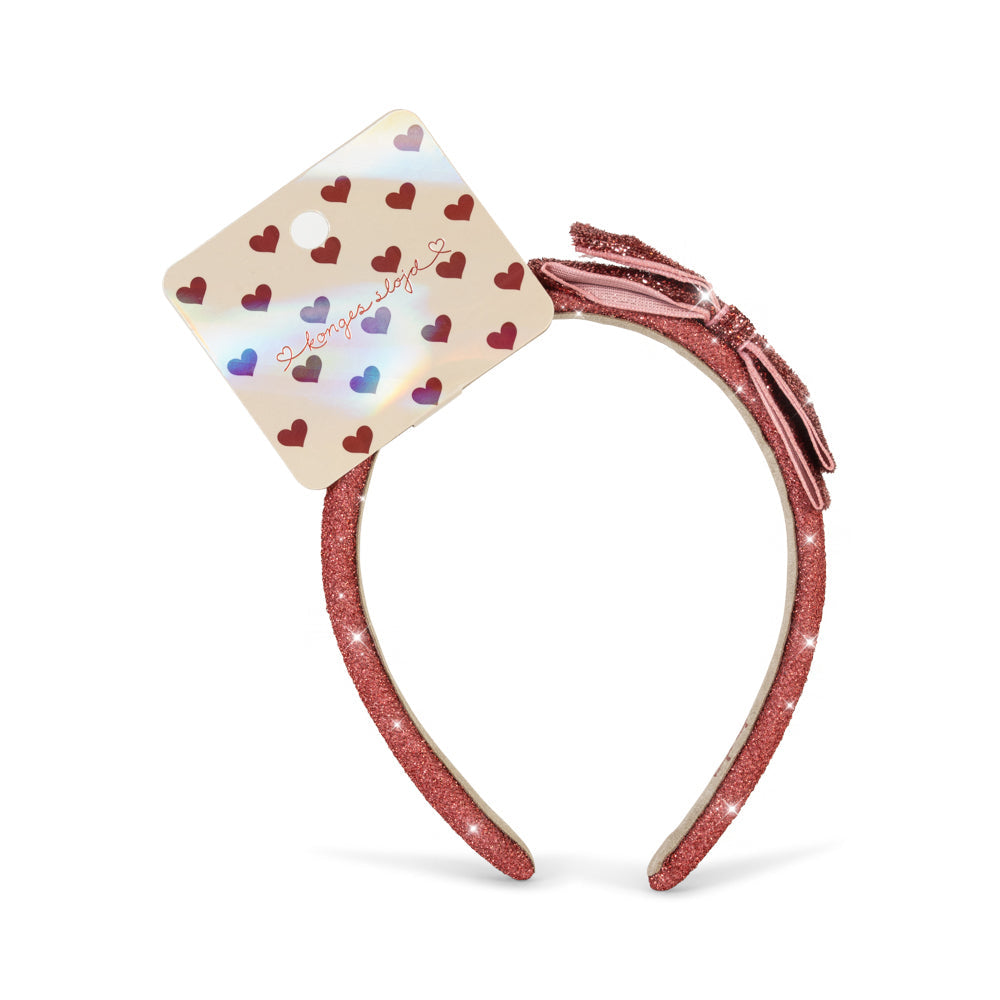 A pink glitter headband featuring a decorative bow on the side and a square card with small red, blue, and purple hearts attached. The card has the word 'Beauty' written on it in cursive, and the headband is photographed on a clean white background.