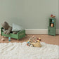 A styled children's bedroom scene with a green wooden bed frame holding a gray stuffed bear and pillows, a pink lamp, a small green cabinet, and a wooden mouse toy driving a car on a white shag rug against a pale green wall.