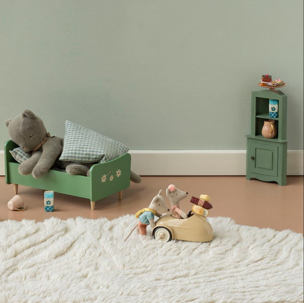 A styled children's bedroom scene with a green wooden bed frame holding a gray stuffed bear and pillows, a pink lamp, a small green cabinet, and a wooden mouse toy driving a car on a white shag rug against a pale green wall.
