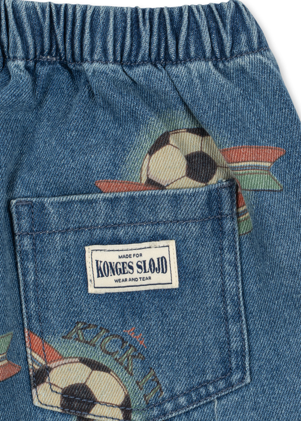Close-up of blue denim shorts with an elastic waistband, a back pocket, and soccer ball prints. A white brand label reading 'KONGES SLOJ' is sewn into the pocket.