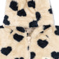 A close-up of a fuzzy hooded jacket in cream with dark navy heart print, featuring a front zipper and a small embroidered brand patch.