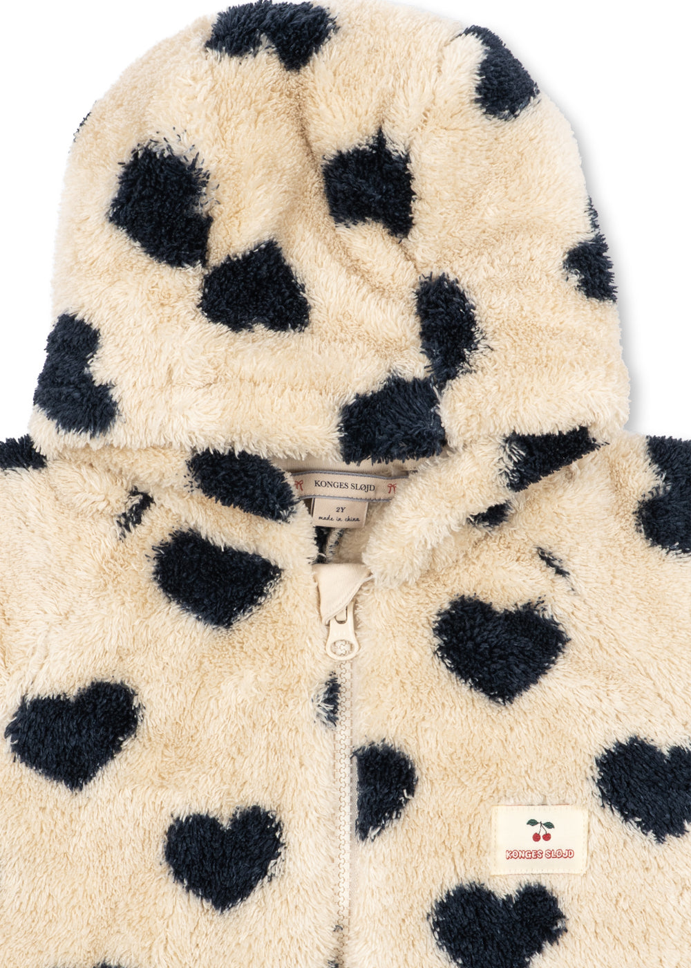 A close-up of a fuzzy hooded jacket in cream with dark navy heart print, featuring a front zipper and a small embroidered brand patch.