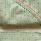 A folded light sage green knit baby garment, likely a wrap or burp cloth, featuring a vertical dotted stripe pattern with small pink hearts and a pink braided trim along the edge.