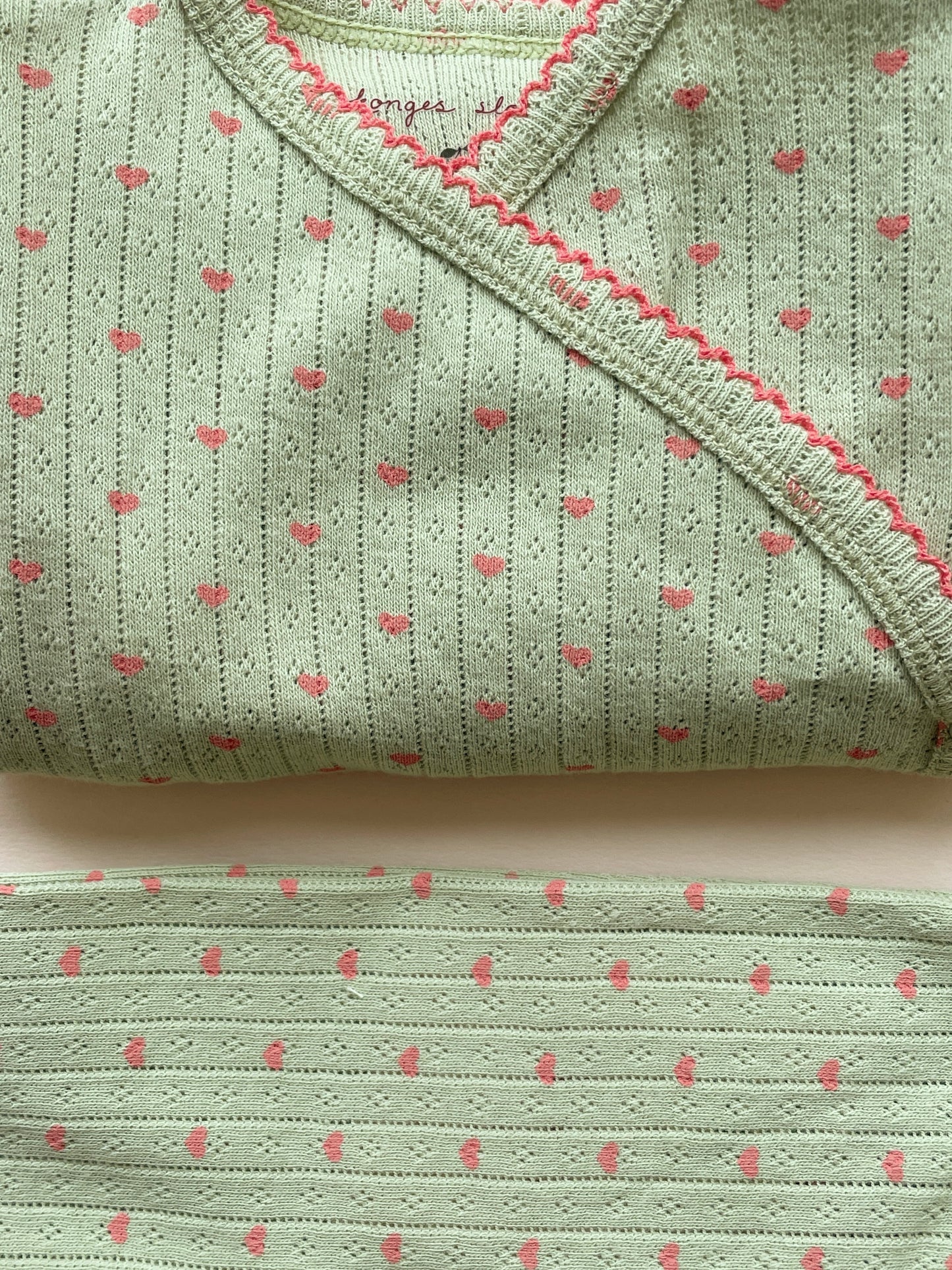A folded light sage green knit baby garment, likely a wrap or burp cloth, featuring a vertical dotted stripe pattern with small pink hearts and a pink braided trim along the edge.