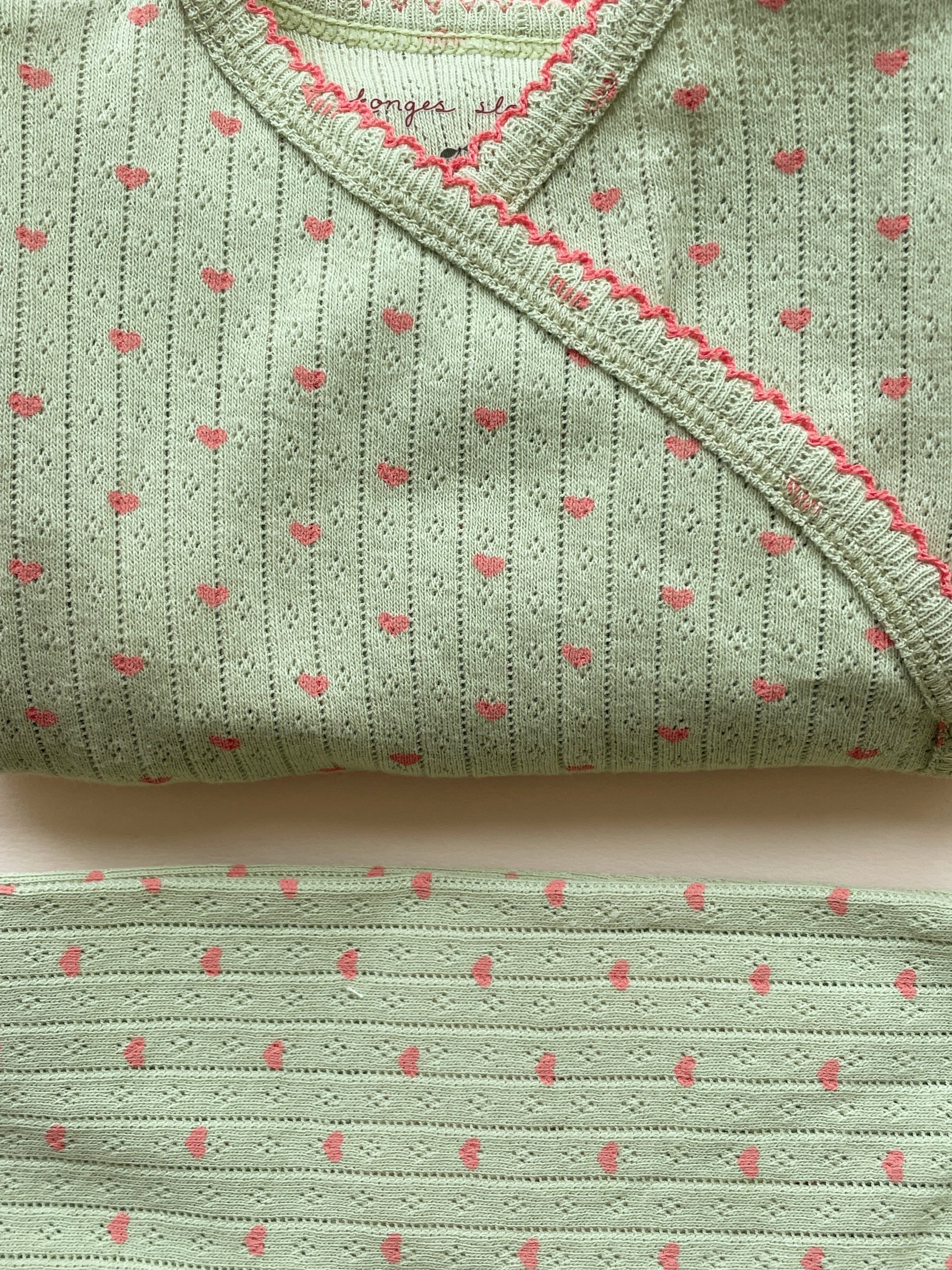 A folded light sage green knit baby garment, likely a wrap or burp cloth, featuring a vertical dotted stripe pattern with small pink hearts and a pink braided trim along the edge.