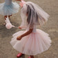 Two young girls in tulle party dresses on a sandy ground. The girl in the foreground wears a light pink tulle dress with a matching veil and pink shoes, while the girl in the background wears a light blue tulle dress with a veil and blue shoes.