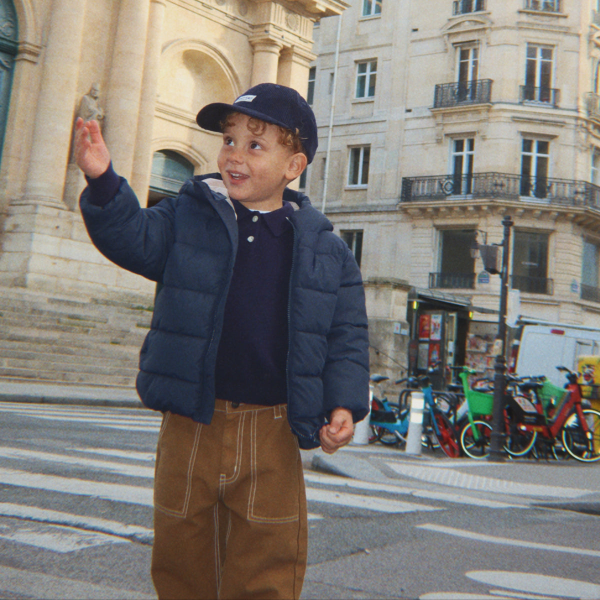 Child in a blue puffer jacket and cap standing on a city street with buildings in the background
