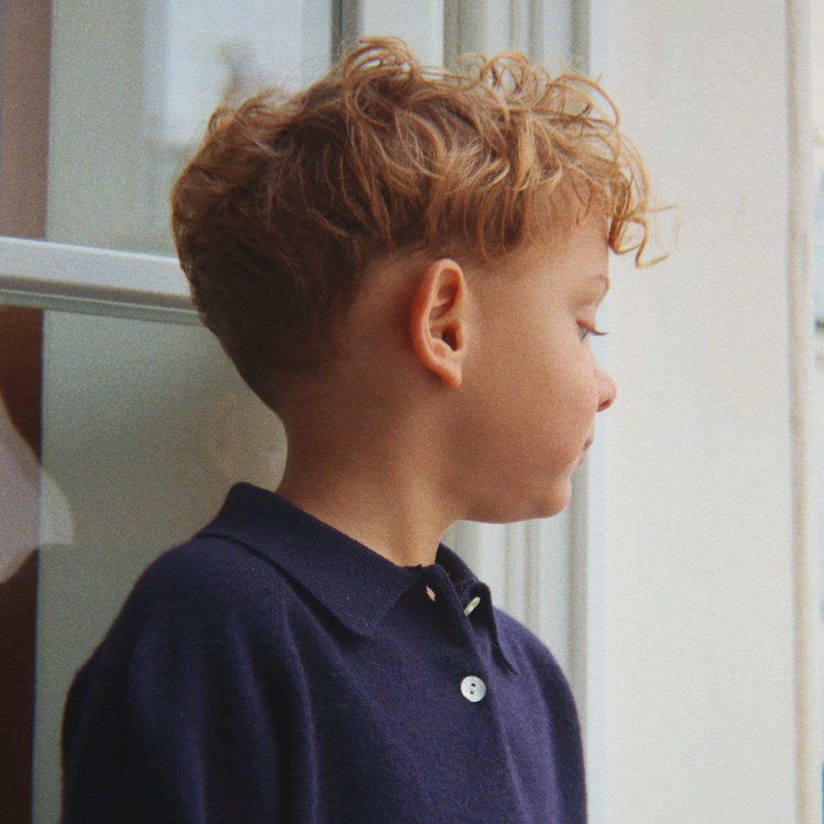 A young boy with curly light brown hair is shown in profile wearing a dark navy blue knit polo sweater. He is standing indoors near a window or door frame.